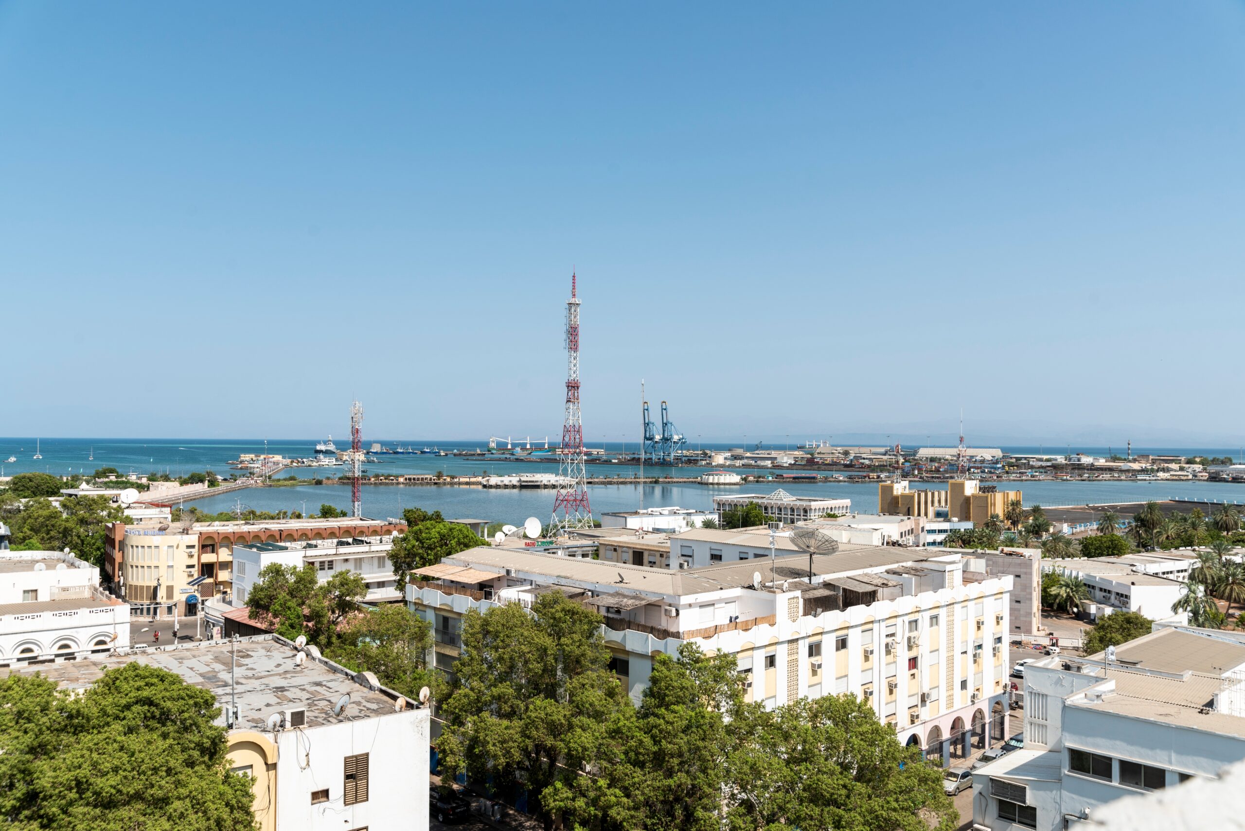 A working port with two cranes sits on the edge of a downtown area with lowrise, white-stone buildings. A lot of green trees line the streets.