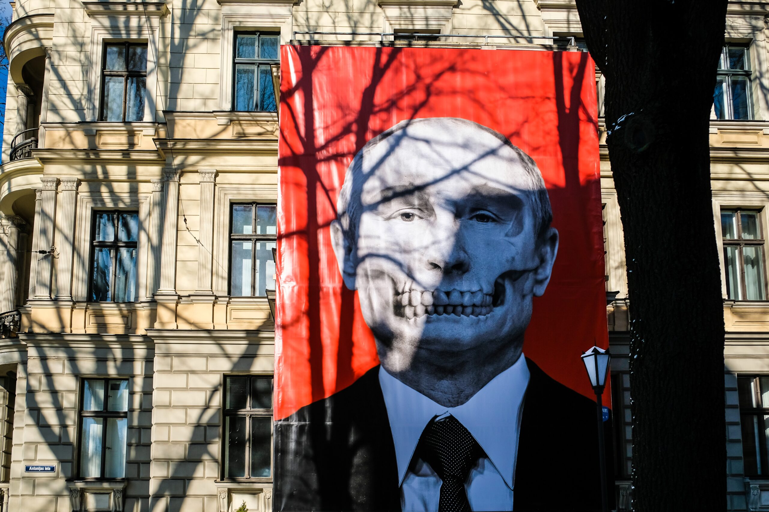 The poster shows Putin’s head and shoulders. He’s in a black suit and tie with a white shirt. Superimposed on his face is a skull, with a faint outline of the sunken bone structure around his eyes. A set of exposed teeth cover Putin’s mouth. The building behind the poster is an elegant sandstone structure with curved balconies with wrought iron railings. Shadows of tree branches cover the building and poster.