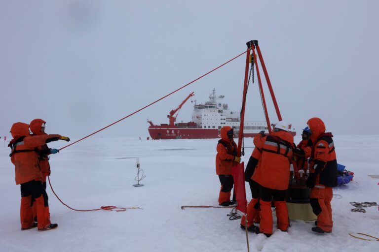 A crew of a half dozen people dressed in red winter gear use a small portable tripod winch system. A red ship is parked in the ice behind them. The sky and surface are a monochromatic white-grey colour where the horizon is barely visible.