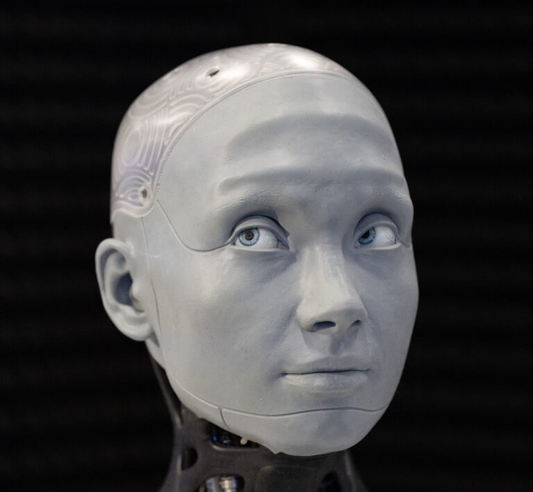 A bald, grey robot-like head with blue eyes.