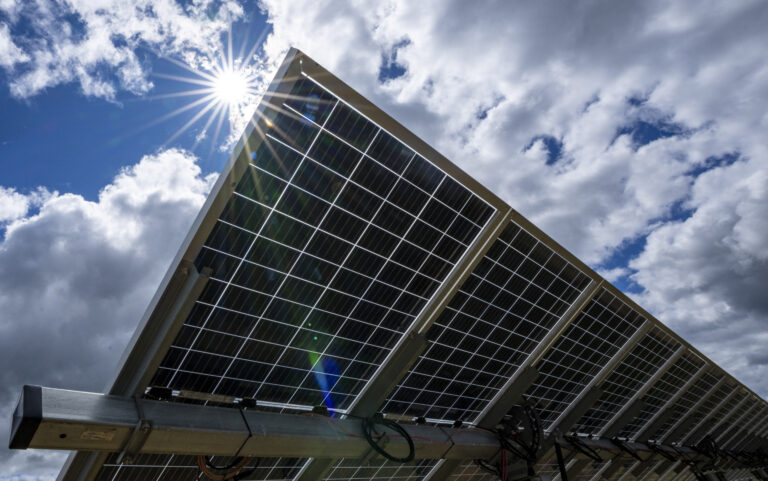 Un employé d’Hydro-Québec vêtu d’un dossard orange et jaune marche devant un énorme panneau solaire. Des nuages planent au-dessus de la scène.