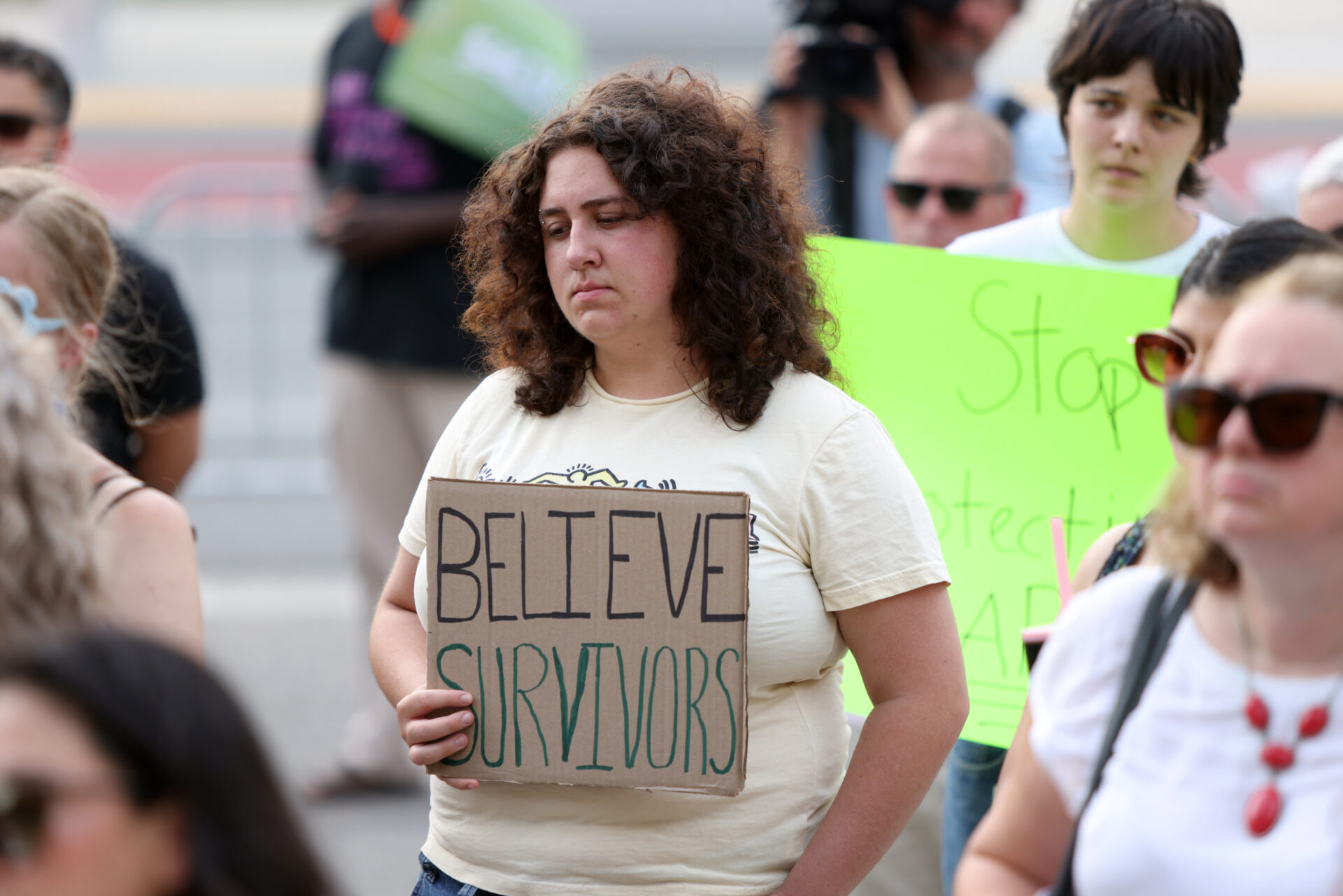 A woman in the middle of a loose crowd is holding a cardboard sign that says “Believe survivors.” She has a solemn expression.
