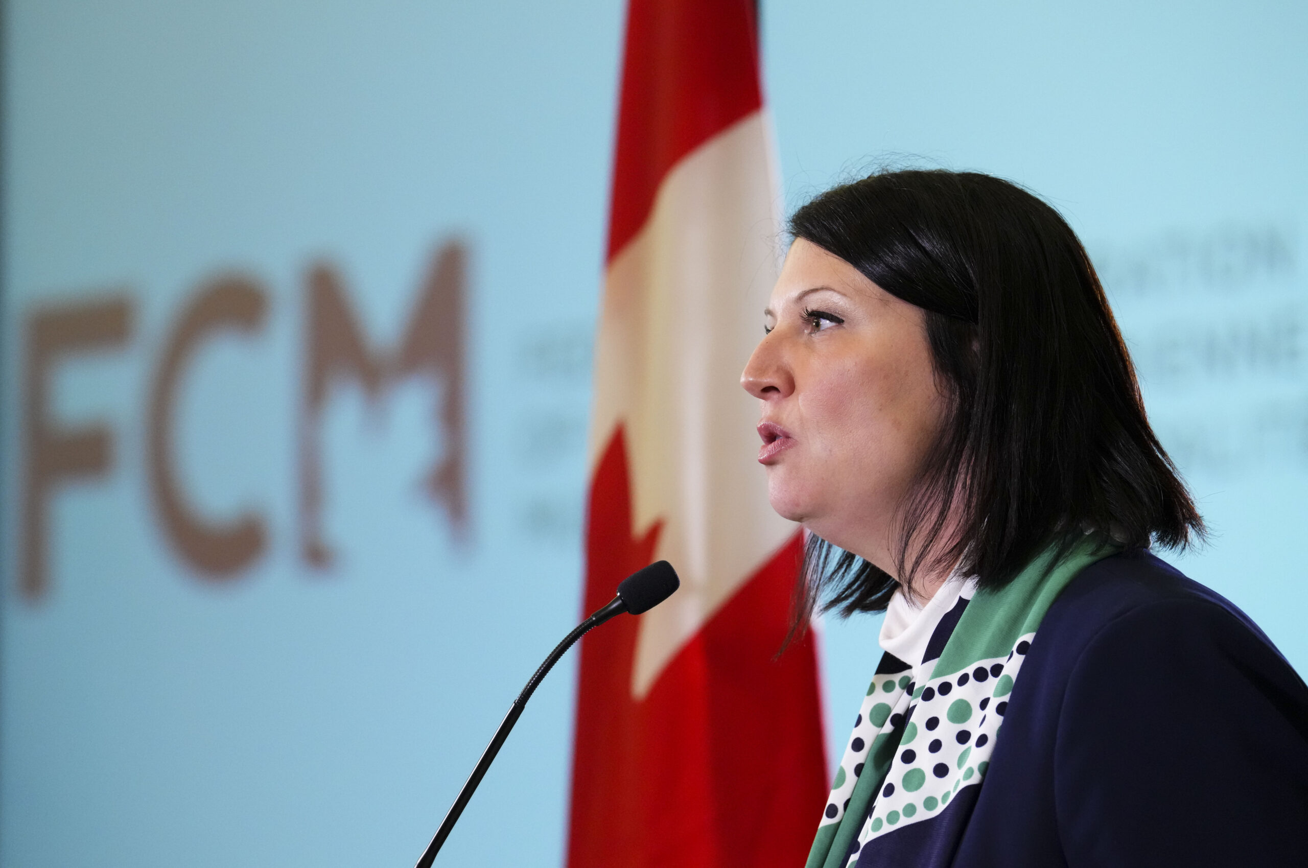 Femme parlant devant un micro, avec le drapeau canadien en arrière-plan et un logo flou de la Fédération canadienne des municipalités.