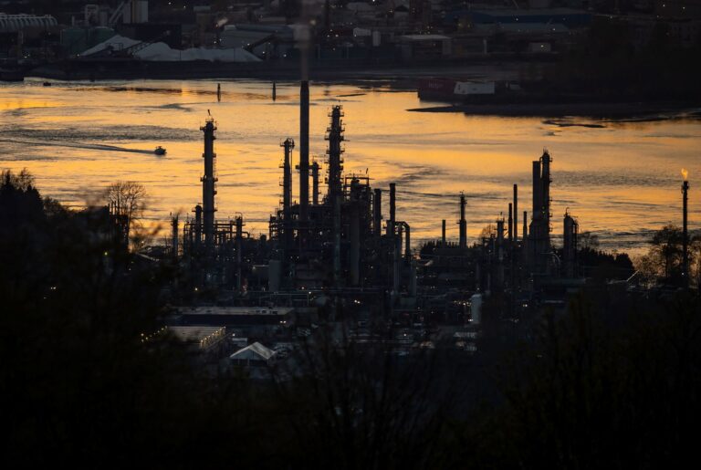 The silhouette of the refinery at sunset with a small boat crossing the orange-glowing waters of the inlet in the background.