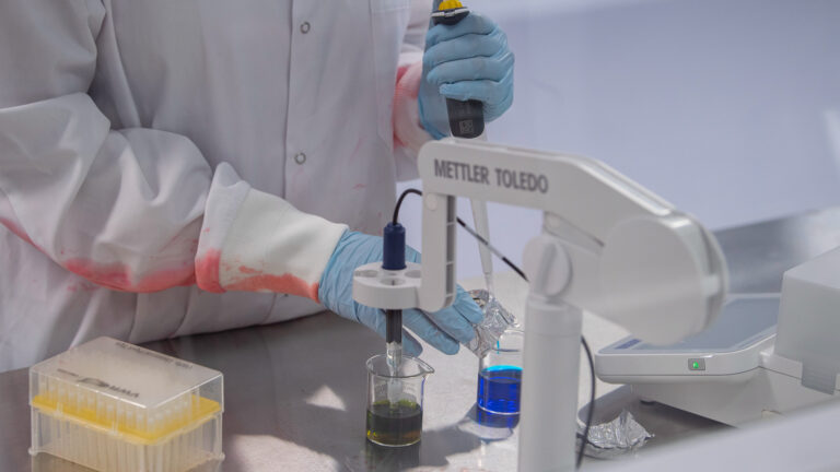 Closeup of the arms of someone in a lab coat and gloves working on a blue sample in a beaker.