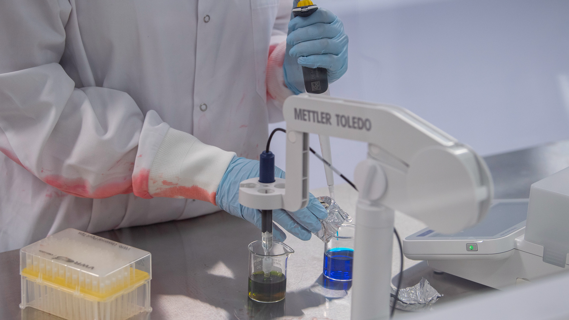 Closeup of the arms of someone in a lab coat and gloves working on a blue sample in a beaker.
