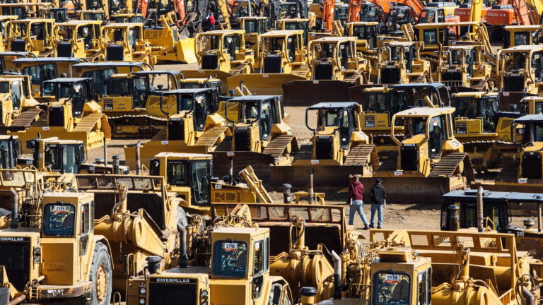 Two men walk in a vast parking lot of bulldozers, excavators, dump trucks and front-end loaders among many other pieces of equipment.