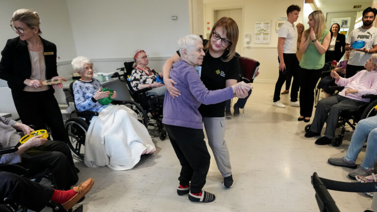 A younger woman dancing with an older woman while several people sit and stand in a circle around them, playing tambourines and other instruments.