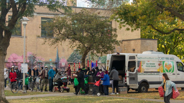A couple dozen people line up behind a white food delivery van. A woman in the foreground walks away with stuffed bags.