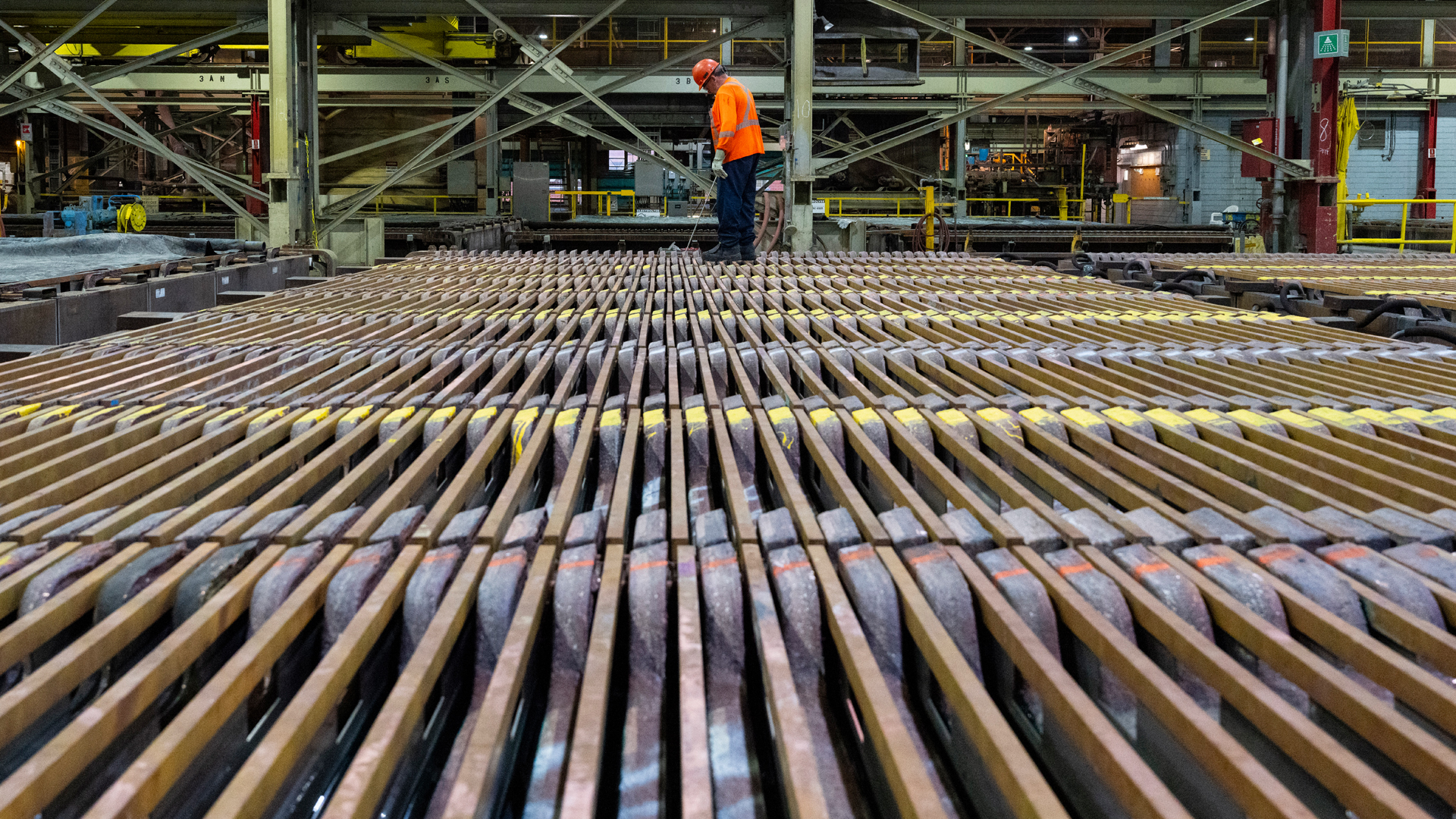 De longues rangées de barres de métal s’étendent à perte de vue dans une usine, avec un seul travailleur portant un gilet orange au loin.