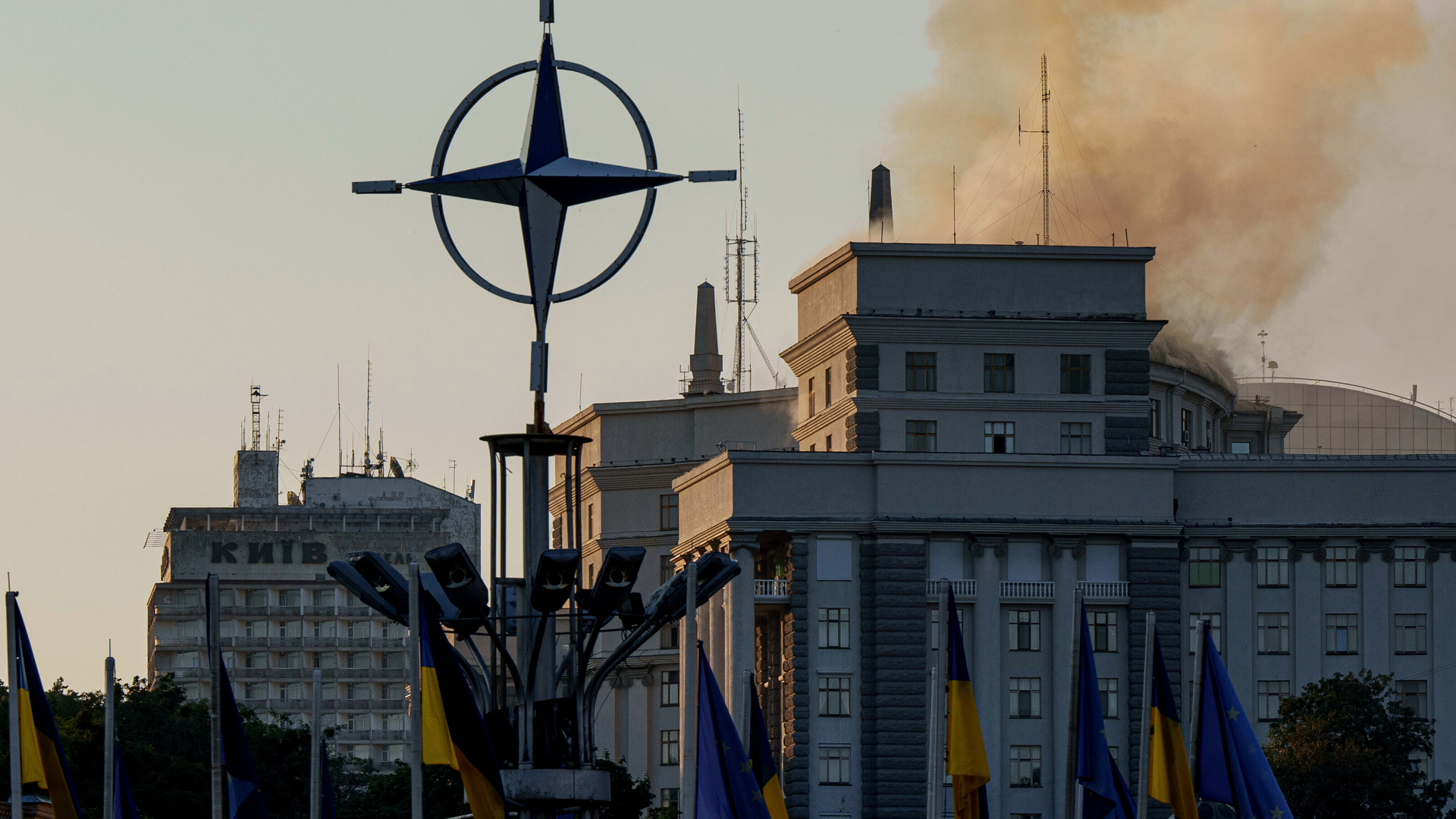 De la fumée s’élève du toit du bâtiment du Cabinet des ministres à Kyiv, en Ukraine. Au premier plan, un monument en forme de rose des vents et plusieurs drapeaux ukrainiens et européens sont visibles.