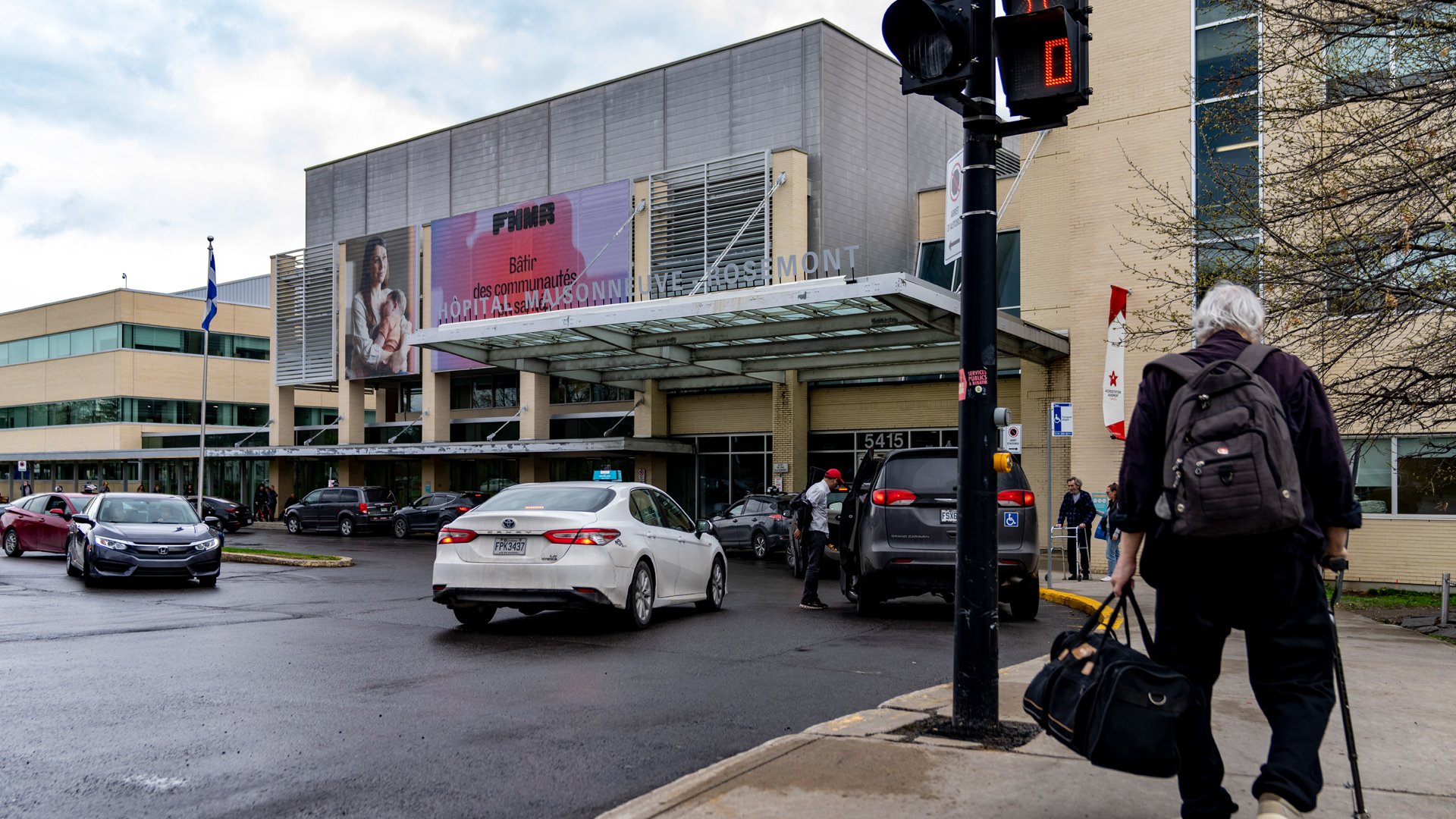 Entrée principale de l’Hôpital Maisonneuve-Rosemont à Montréal, avec des voitures devant le pavillon et un homme marchant avec une canne et un sac de voyage, vu de dos.