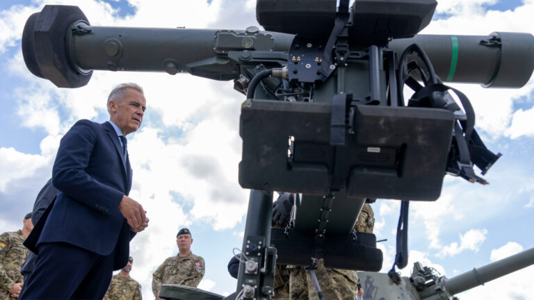 Mark Carney walking beneauth the barrel of a launch tube with soldiers in the background.