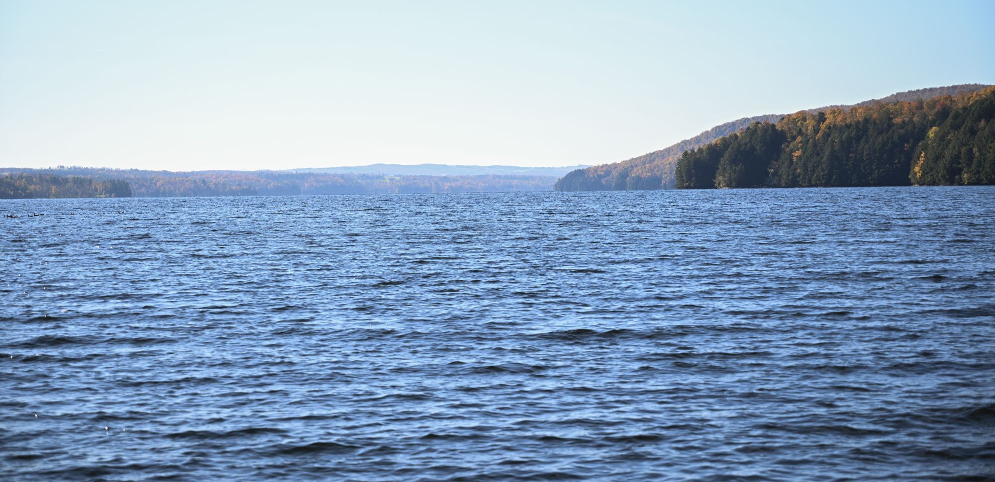 A lake with rolling hills in the background on a sunny day. The leaves are just starting to change colours on the trees.