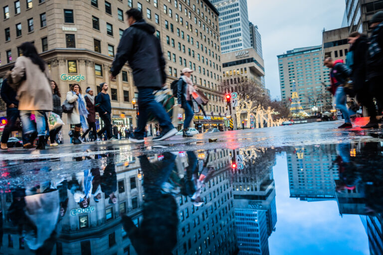 Des passants traversent une intersection animée au centre-ville, sous un ciel gris. Les immeubles environnants, dont un avec le logo de Google, se reflètent dans une flaque d’eau au premier plan. Les décorations lumineuses des arbres évoquent la période des Fêtes.