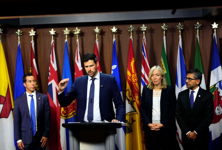 All four are standing in front of a wall lined with provincial flags. Fraser is at a podium.