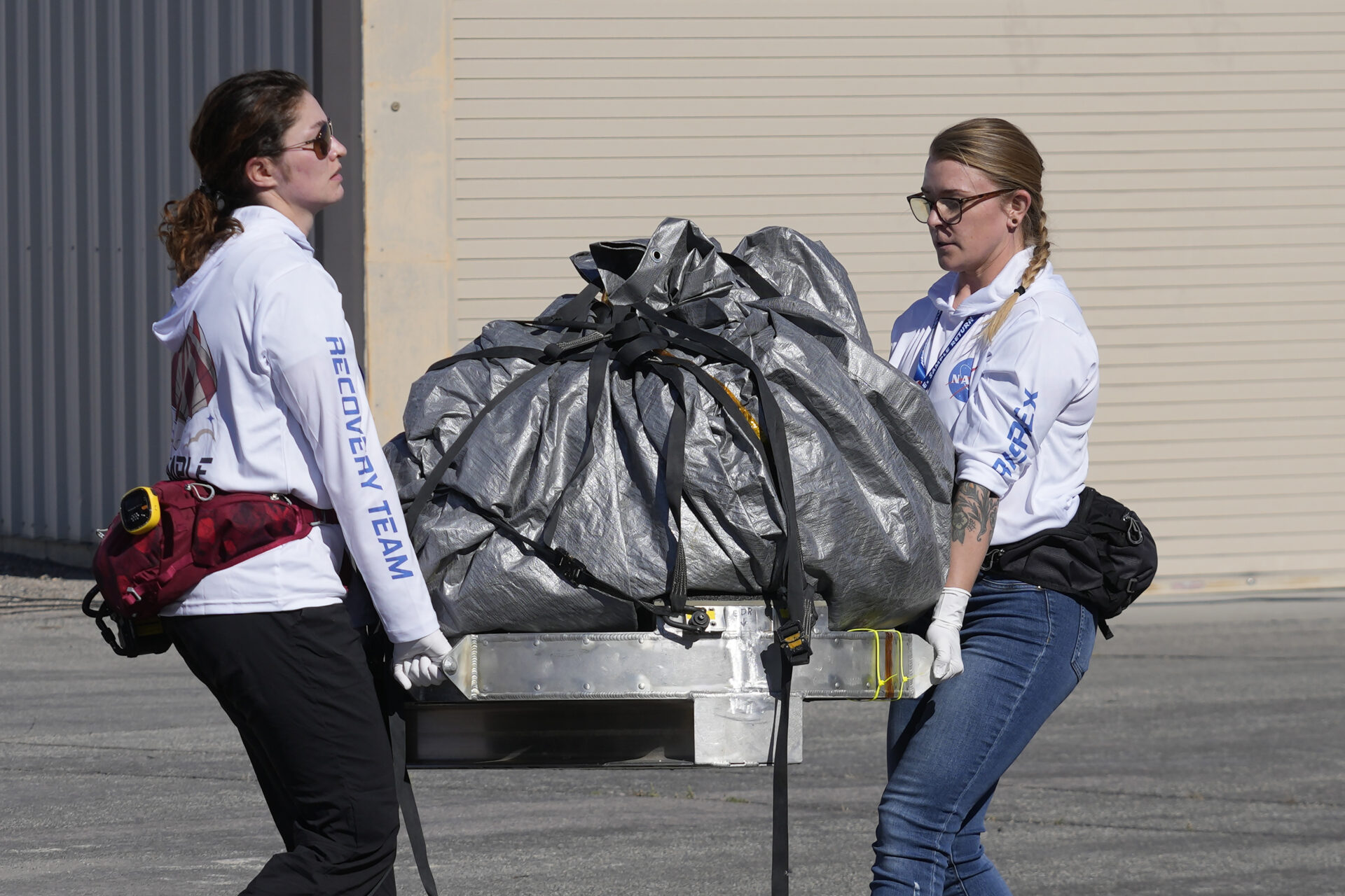 The sample is wrapped up in a grey tarp cinched with a black cords. It’s sitting on a thick metal base with handles on each side, which two white women use to move it.