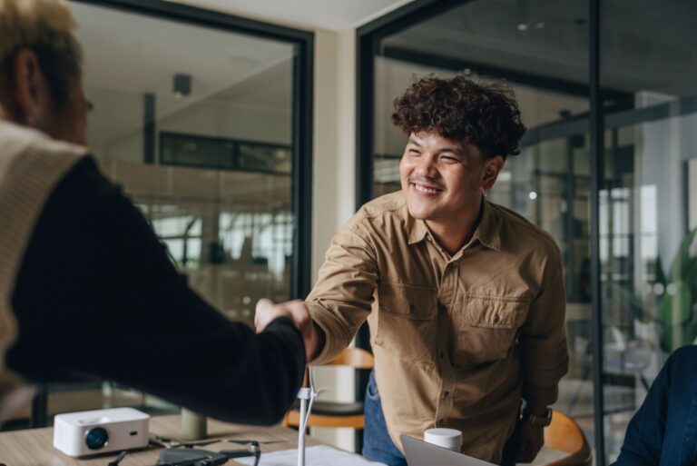 A 20-something man reaches across a table in an office to shake hands with a colleague.