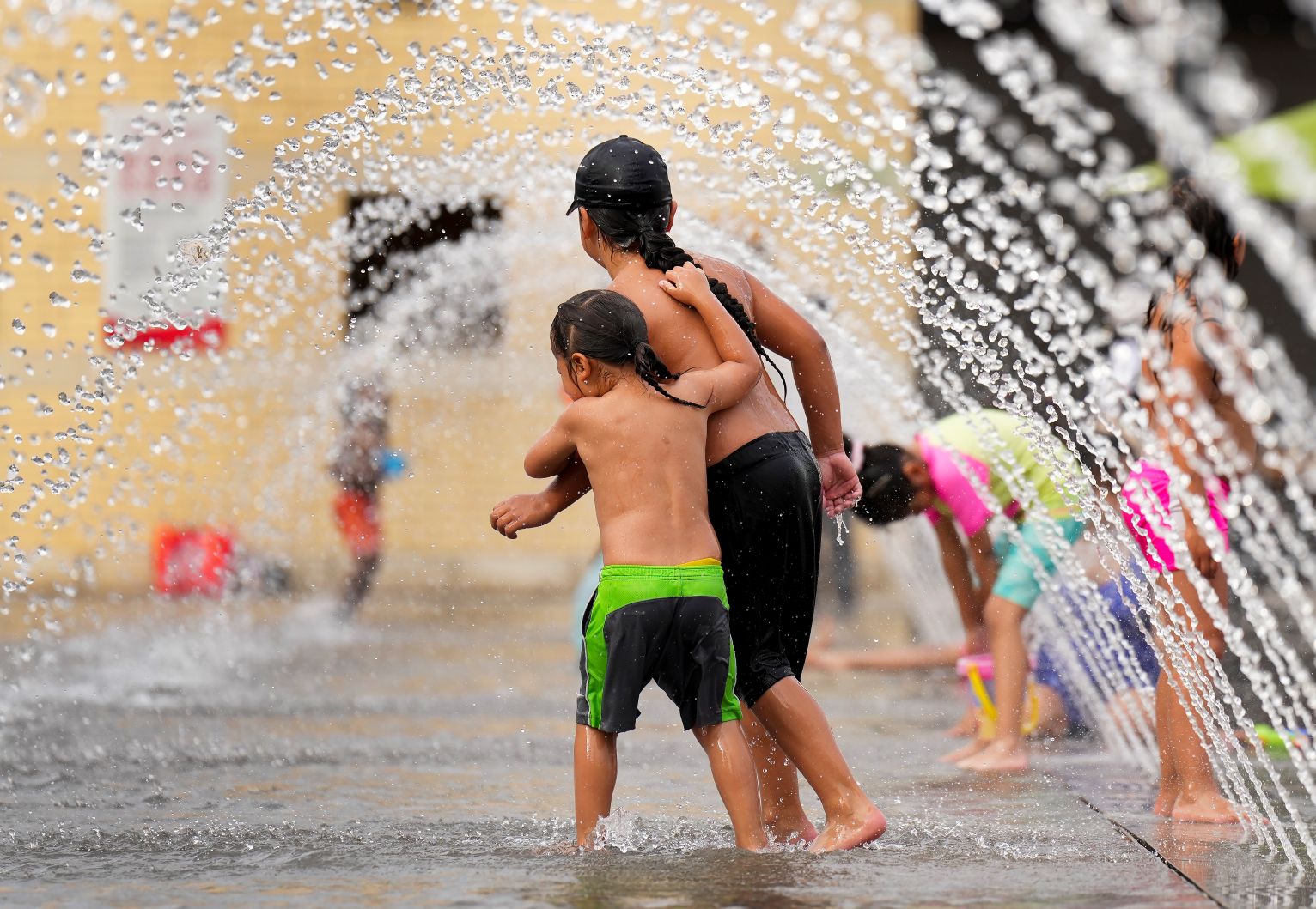Two small children tussle under an arc of water.