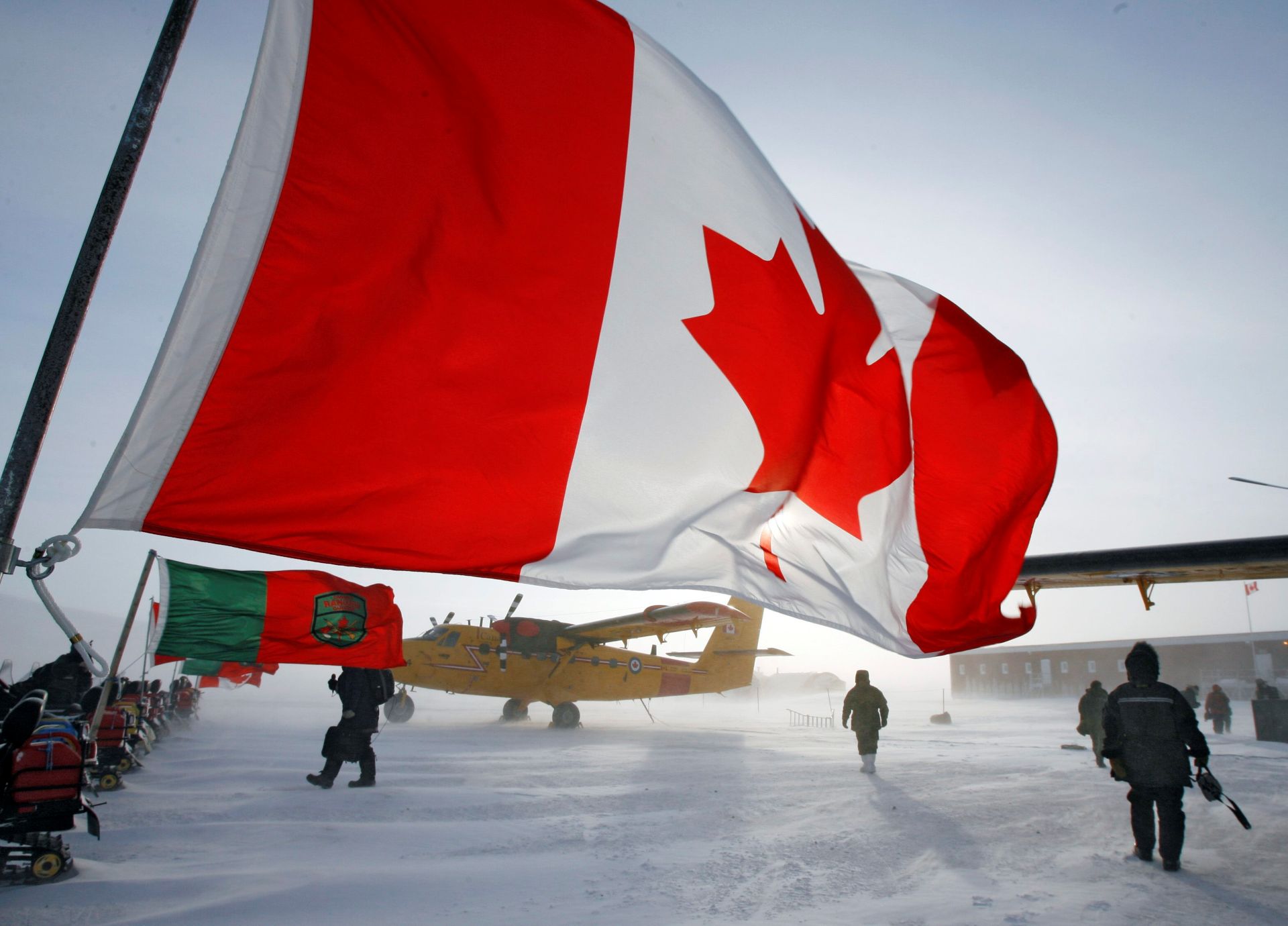 A Canadian flag flies from a row of snowmobiles with a small yellow plane parked nearby. All around is blowing snow on ice.