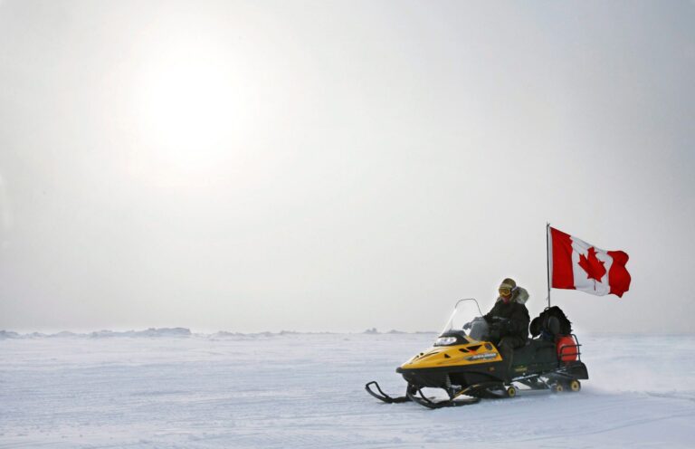 The man is driving a snowmobile across a flat expanse of white, just him, snow, ice and a white sky.