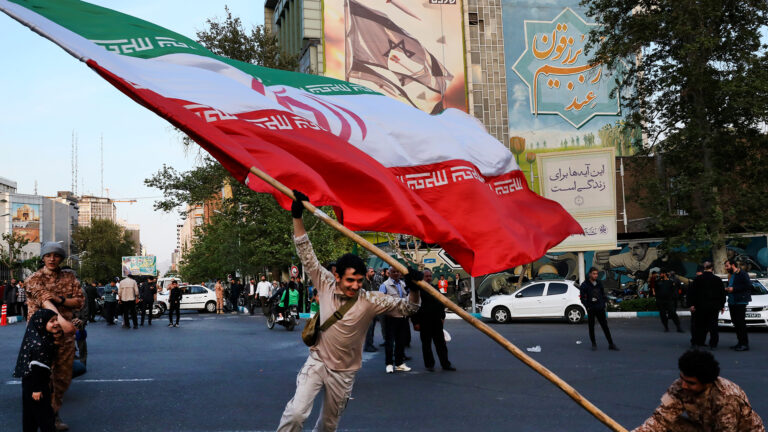 Two men wave a huge Iranian flag in a paved area in front of the building.