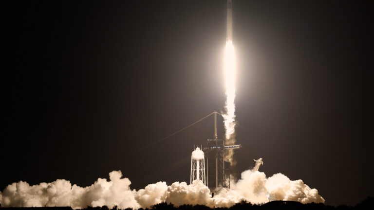 Against a black sky, the white rocket sends blindingly white exhaust gases to the ground as it lifts off.