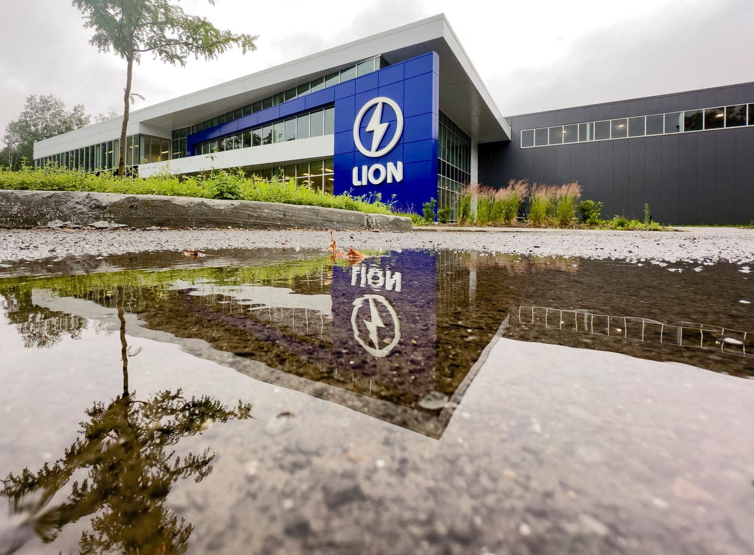 Une flaque d'eau dans un stationnement vide au premier plan, avec une façade de bâtiment flambant neuve sur laquelle sont inscrits « Lion » avec un éclair électrique en blanc.