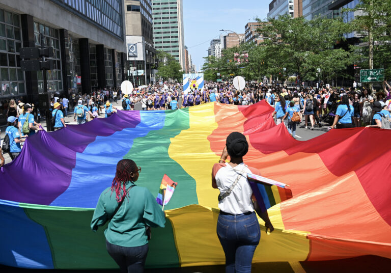 People hold a huge rainbow flag in downtown Montreal during the city’s Pride parade. Clothed in a rainbow of colours under a sunny sky.