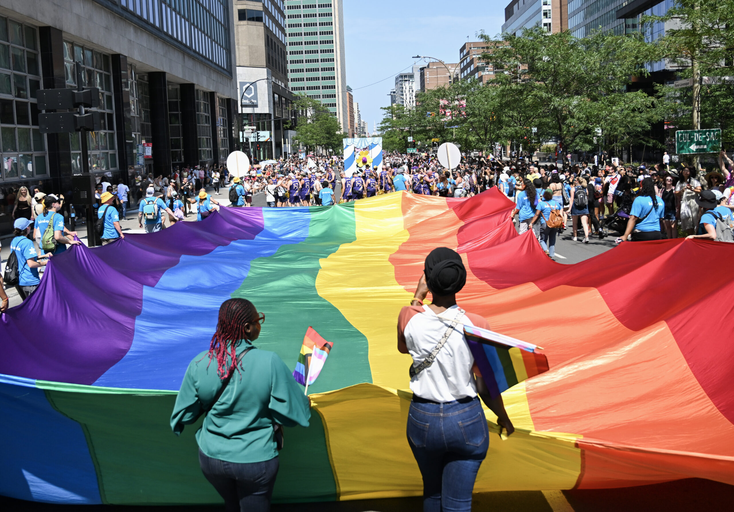 Pride Parade, Des personnes tiennent un immense drapeau arc-en-ciel déployé dans une rue du centre-ville de Montréal pendant le défilé de la Fierté. La foule, vêtue de couleurs vives, borde la rue sous un ciel ensoleillé.