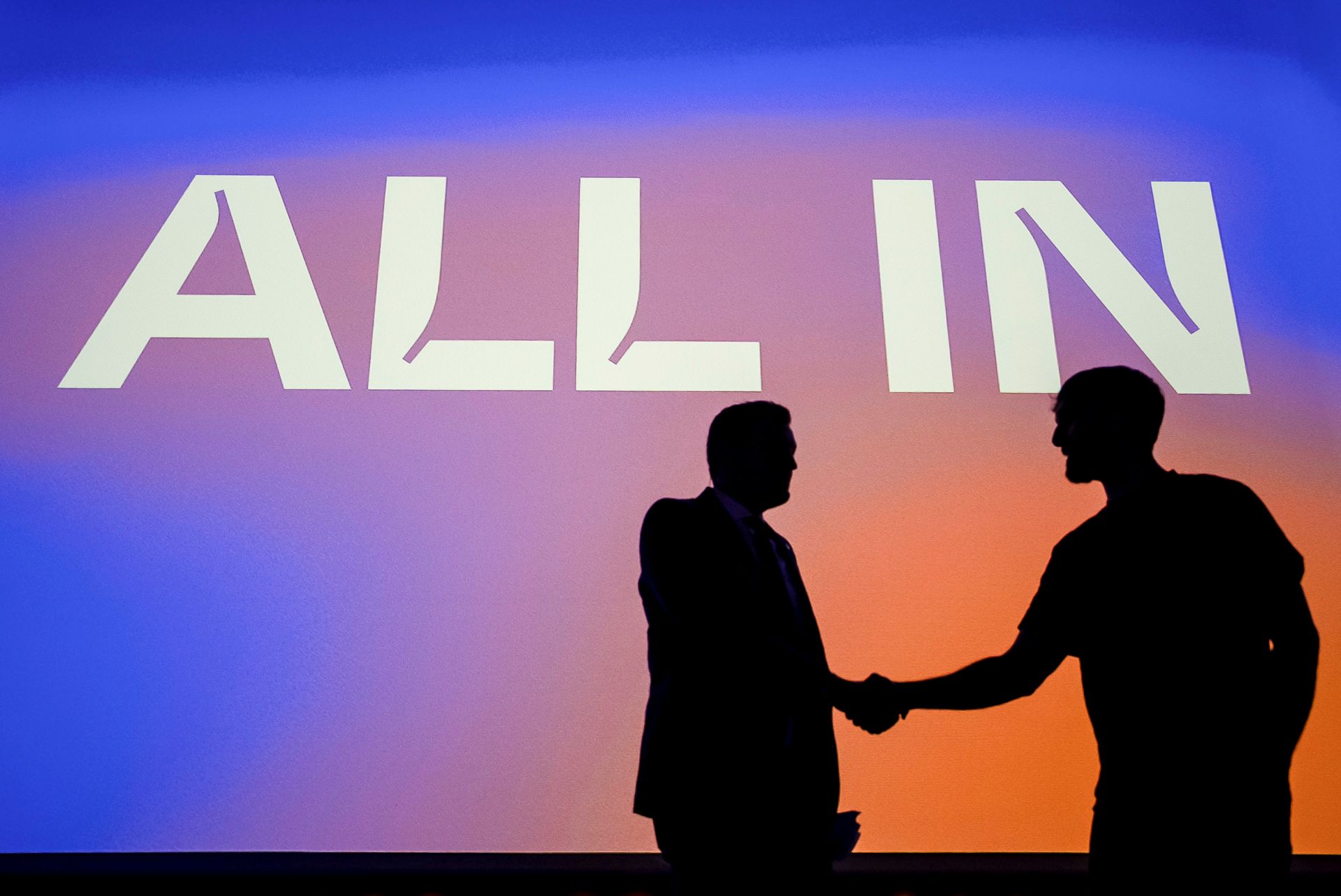 The two men are in silhouette against a large screen that says “All In.” The screen is blue and mauve.