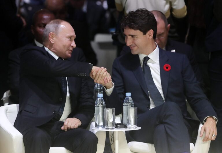 Putin and Trudeau sitting in easy chairs with strained smiles as they shake hands.