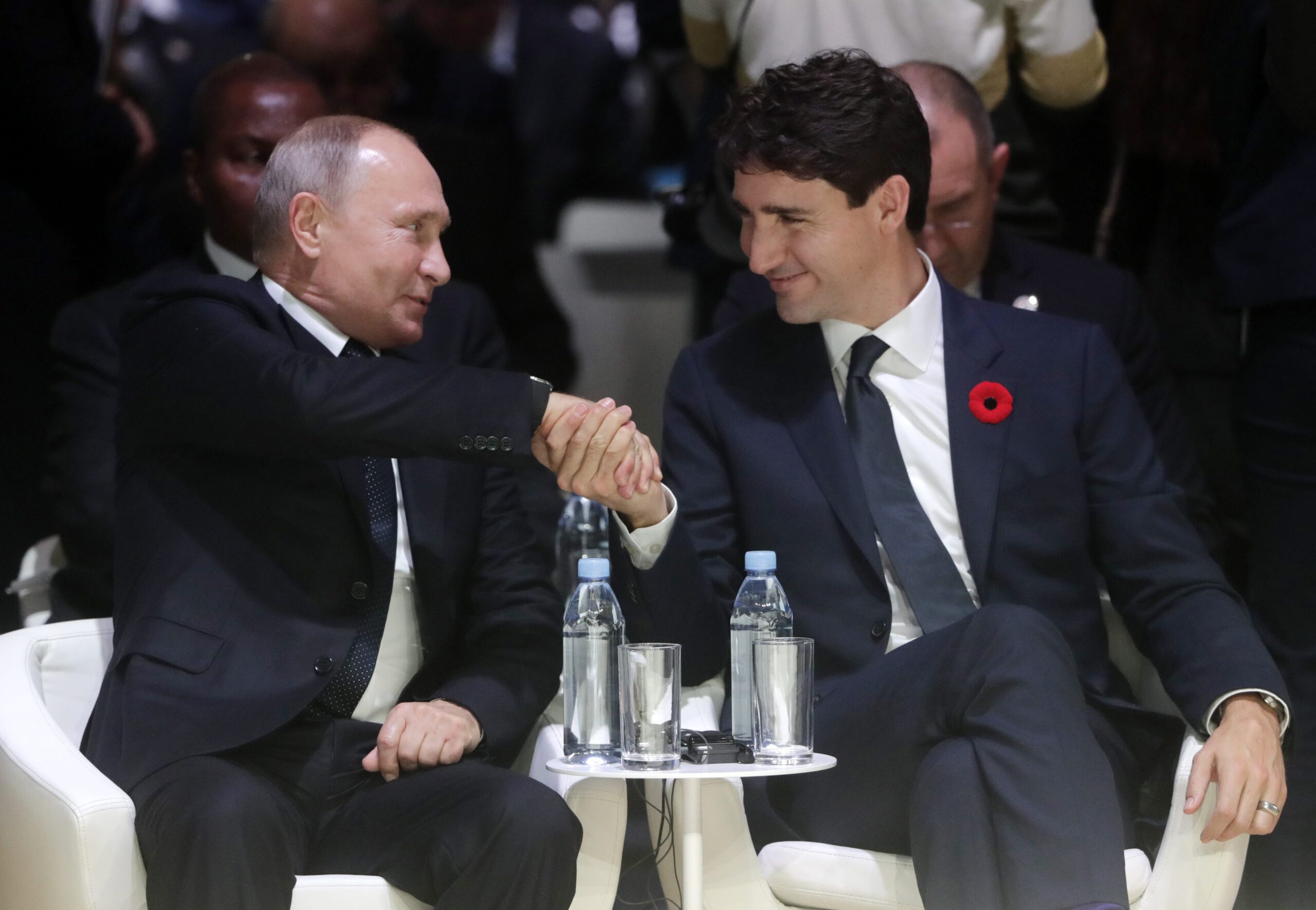 Trudeau et Poutine sont assis de chaque côté d’une petite table ronde. Trudeau serre la main de Poutine. Des bouteilles d’eau sont déposées sur la table,