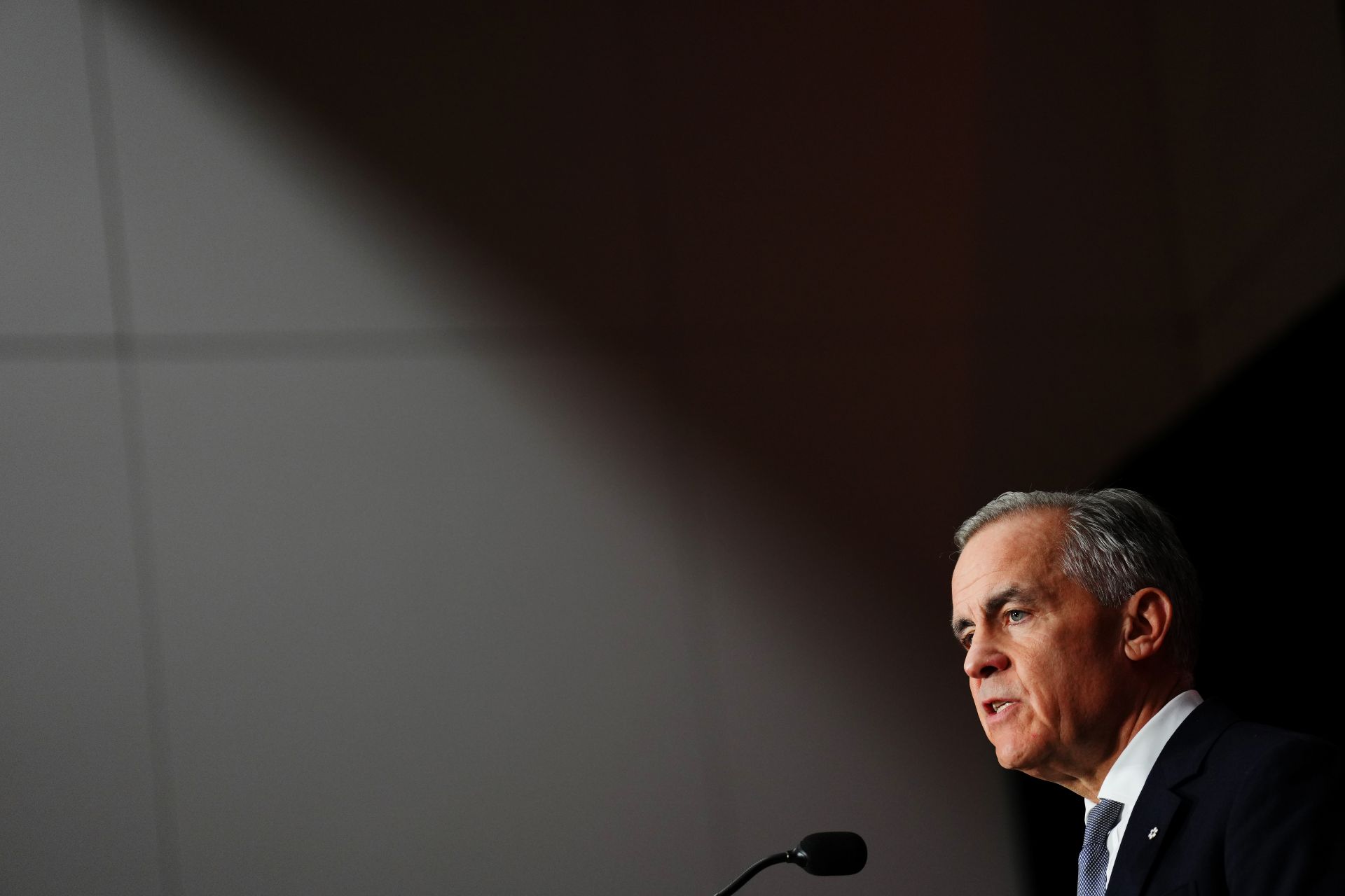 Mark Carney speaking at a podium, viewed from the side, with dark background.