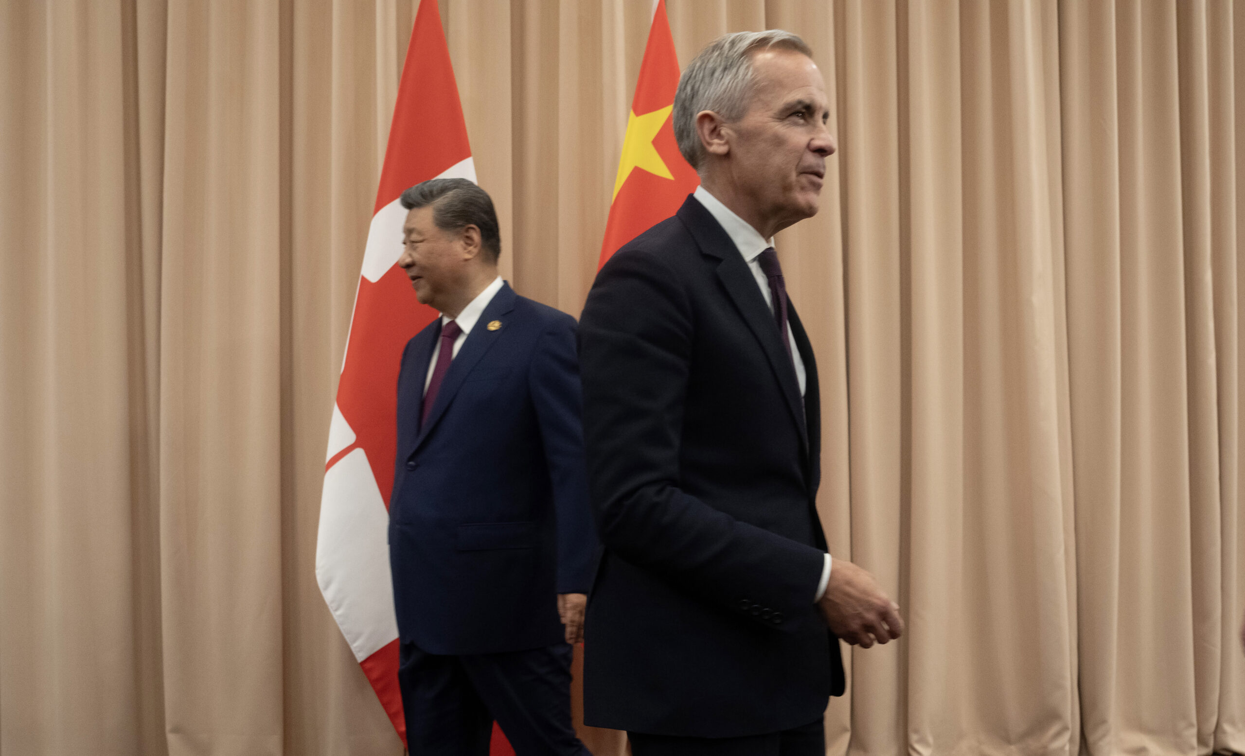 Mark Carney et Xi Jinping en complet noir se tiennent de dos l’un et l’autre devant des drapeaux du Canada et de la Chine, dans une salle aux rideaux beiges.