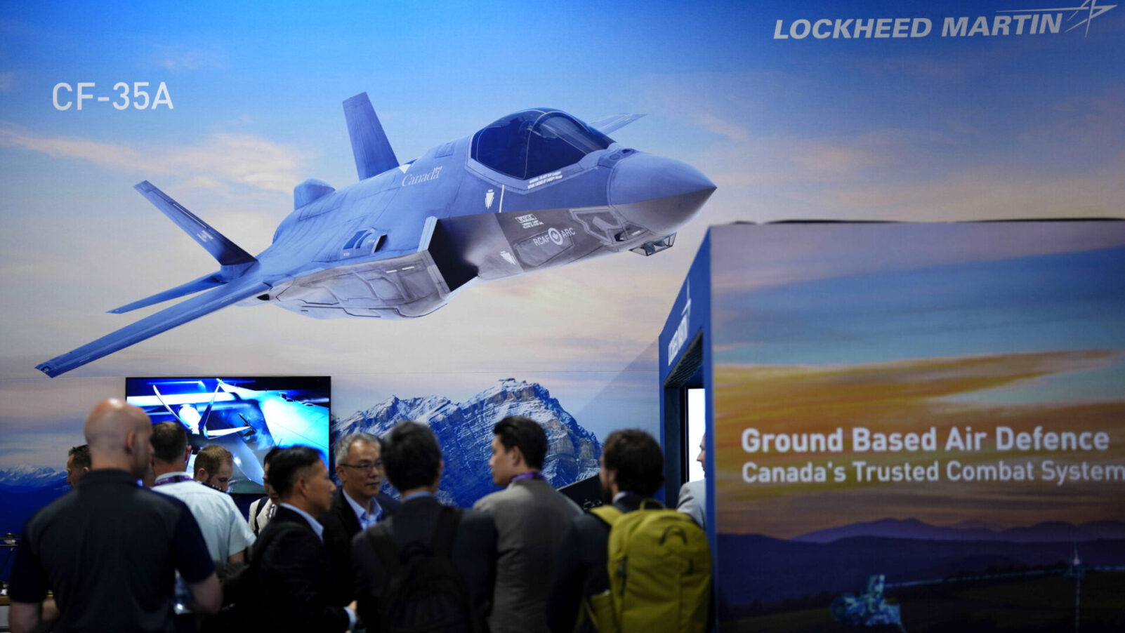 L'image d'un CF-35 est exposée au kiosque de Lockheed Martin lors du salon annuel CANSEC de l’Association canadienne de l’industrie de la défense et de la sécurité, à Ottawa. L’avion est présenté de profil. Autour de lui, plusieurs visiteurs observent et discutent.