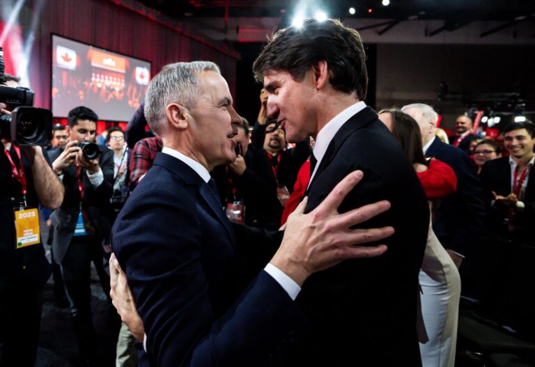 Mark Carney holds Justin Trudeau by the biceps while they are surrounded by photographers and party members.