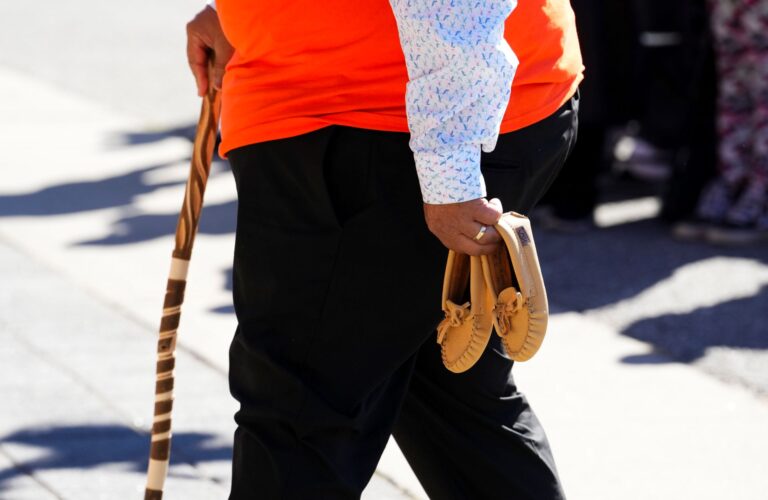 In one hand is the pair of tan moccasins. With his other hand he holds a wooden cane. He’s wearing an orange t-shirt over a long-sleeved shirt.