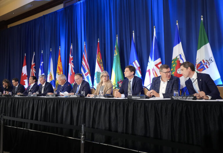 The premiers are seated behind a table on a stage. They are lined up in front of a series of flags representing Canada’s provinces and territories, set against a blue curtain.
