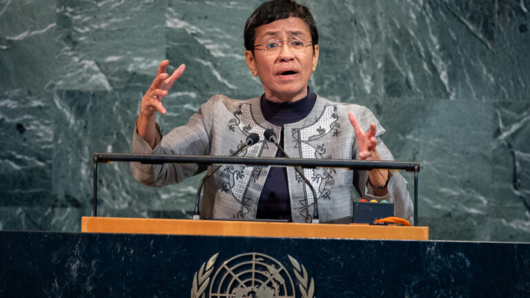 Maria Ressa gestures with both hands from the behind the podium of the general assembly with the UN logo on it.