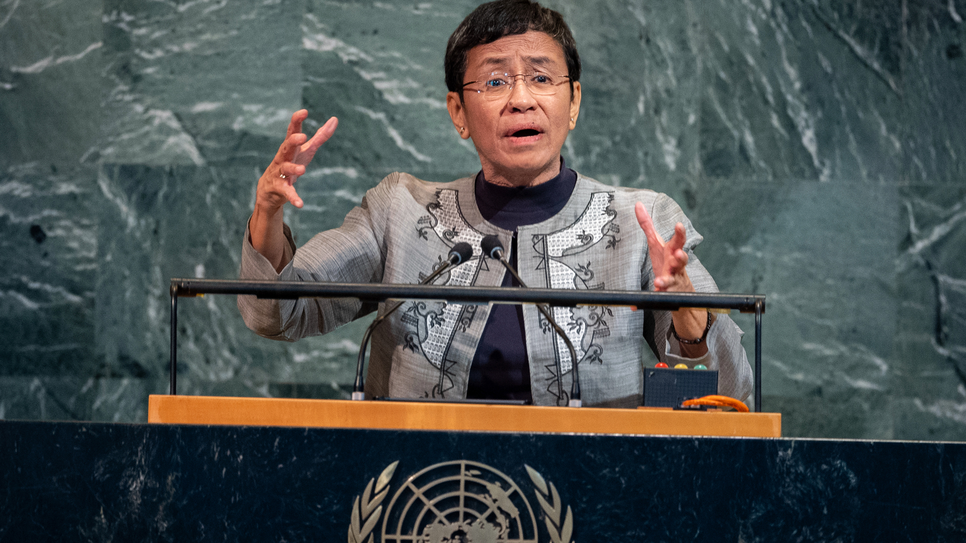 Maria Ressa gestures with both hands from the behind the podium of the general assembly with the UN logo on it.
