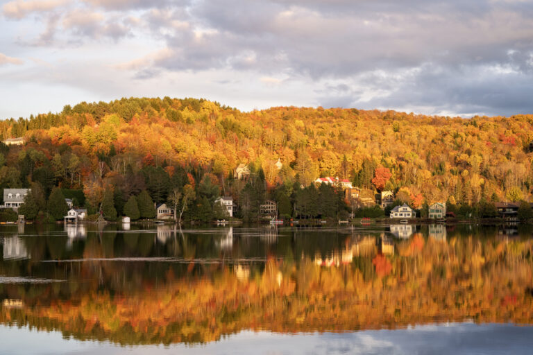 Lac calme bordé de maisons et de chalets, avec la forêt en pleine couleurs d’automne se reflétant dans l’eau, sous un ciel partiellement nuageux.