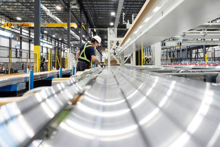 A worker in a hardhat wearing in a black T-shirt with a yellow reflective vest inspects a long, curved piece of aluminum.