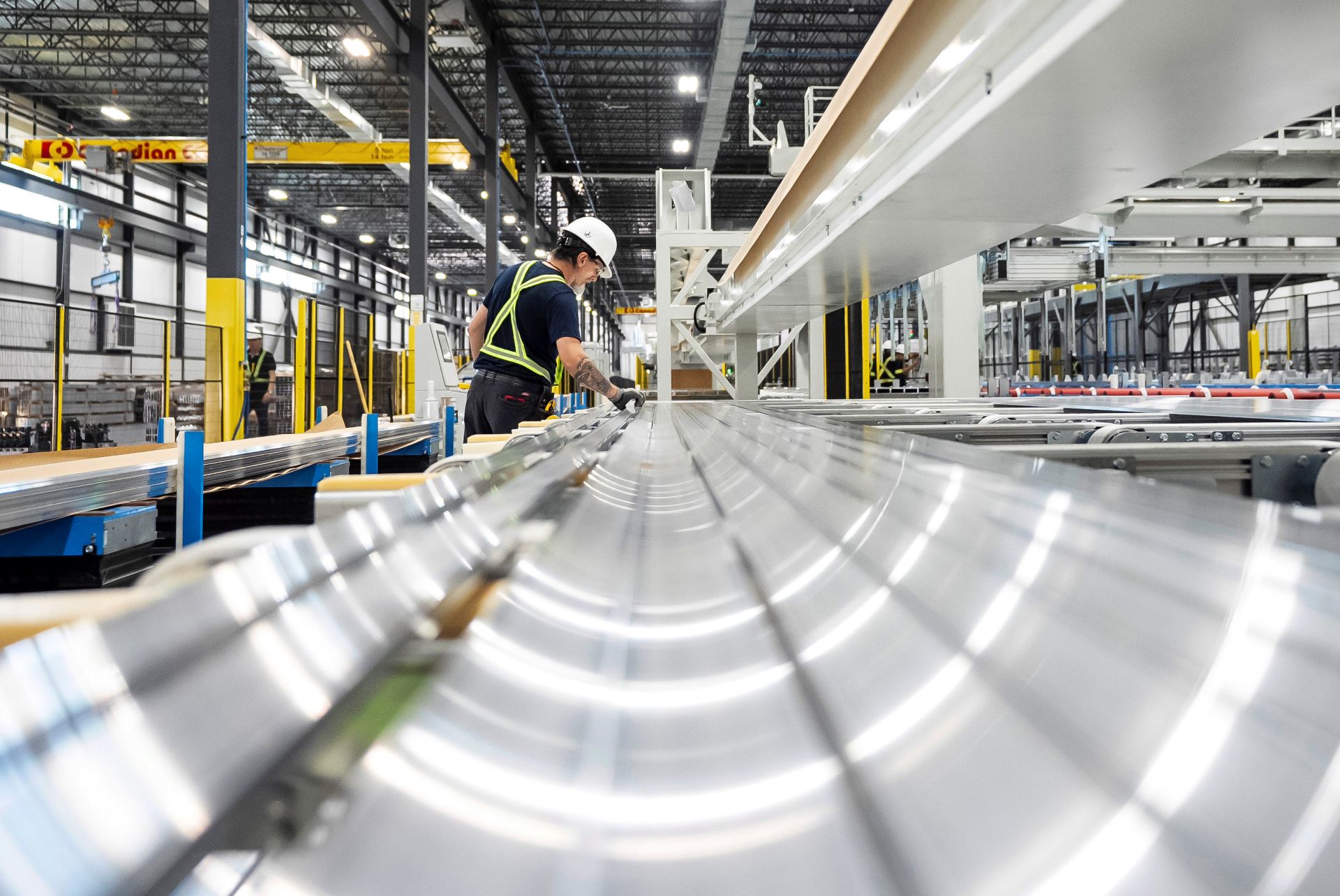 A worker in a hardhat wearing in a black T-shirt with a yellow reflective vest inspects a long, curved piece of aluminum.