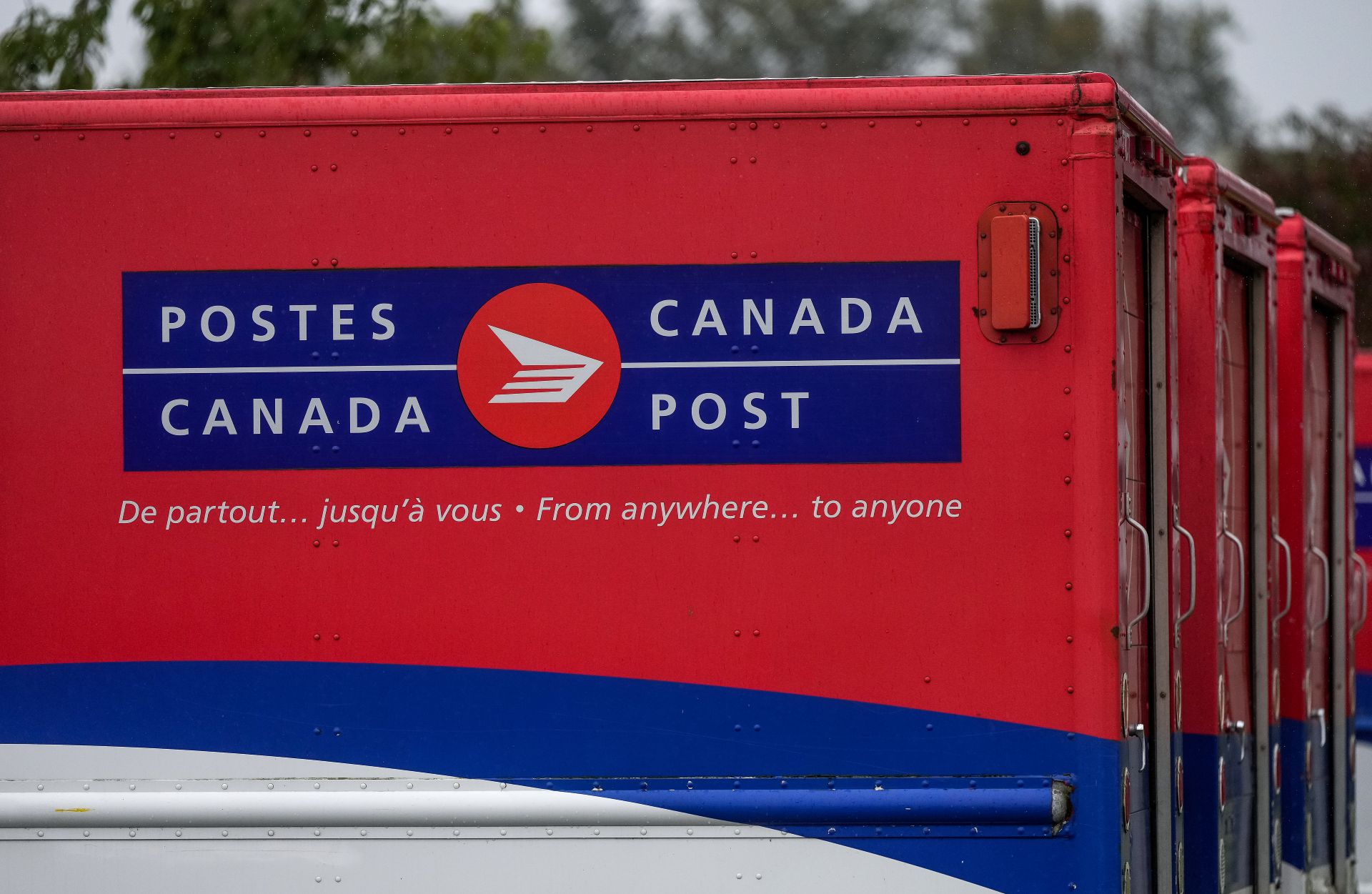 A line of Canada Post red, blue and white trucks with the slogan “de partout . . . jusqu’à vous” and “From anywhere . . . to anyone” written on the side.