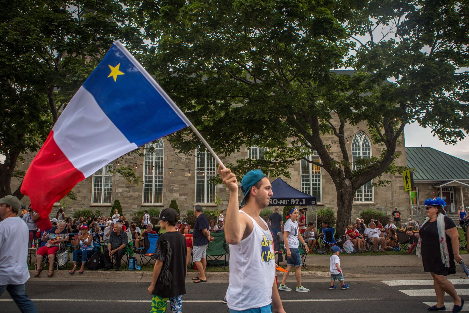 A man in a whit shirt and ballcap waves a large Acadian flag as he walks.