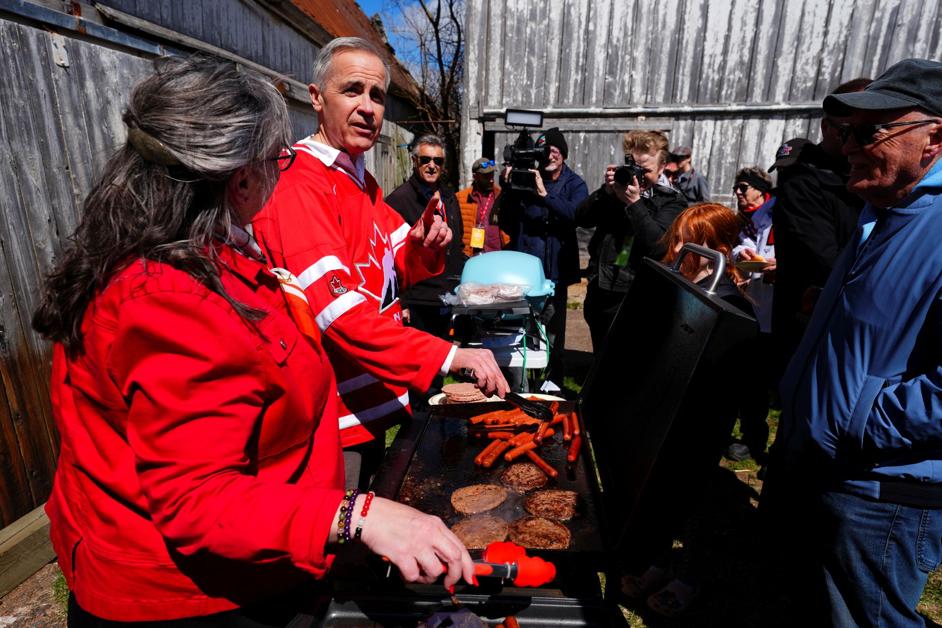 Mark Carney wearing a Team Canada sweater while working the BBQ with some supporters.