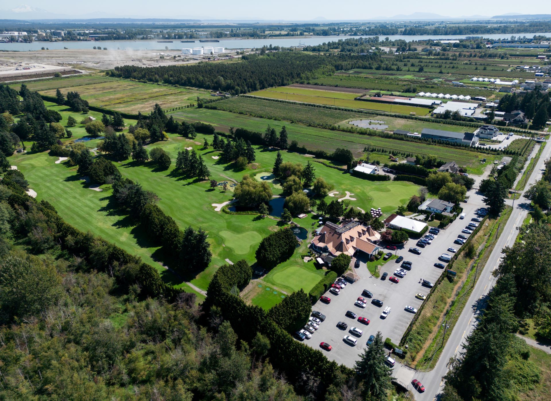 Aerial view of the club and its green expanse dotted with evergreens. Adjacent to it are strips of farm land and a cleared industrial area near a body of water.
