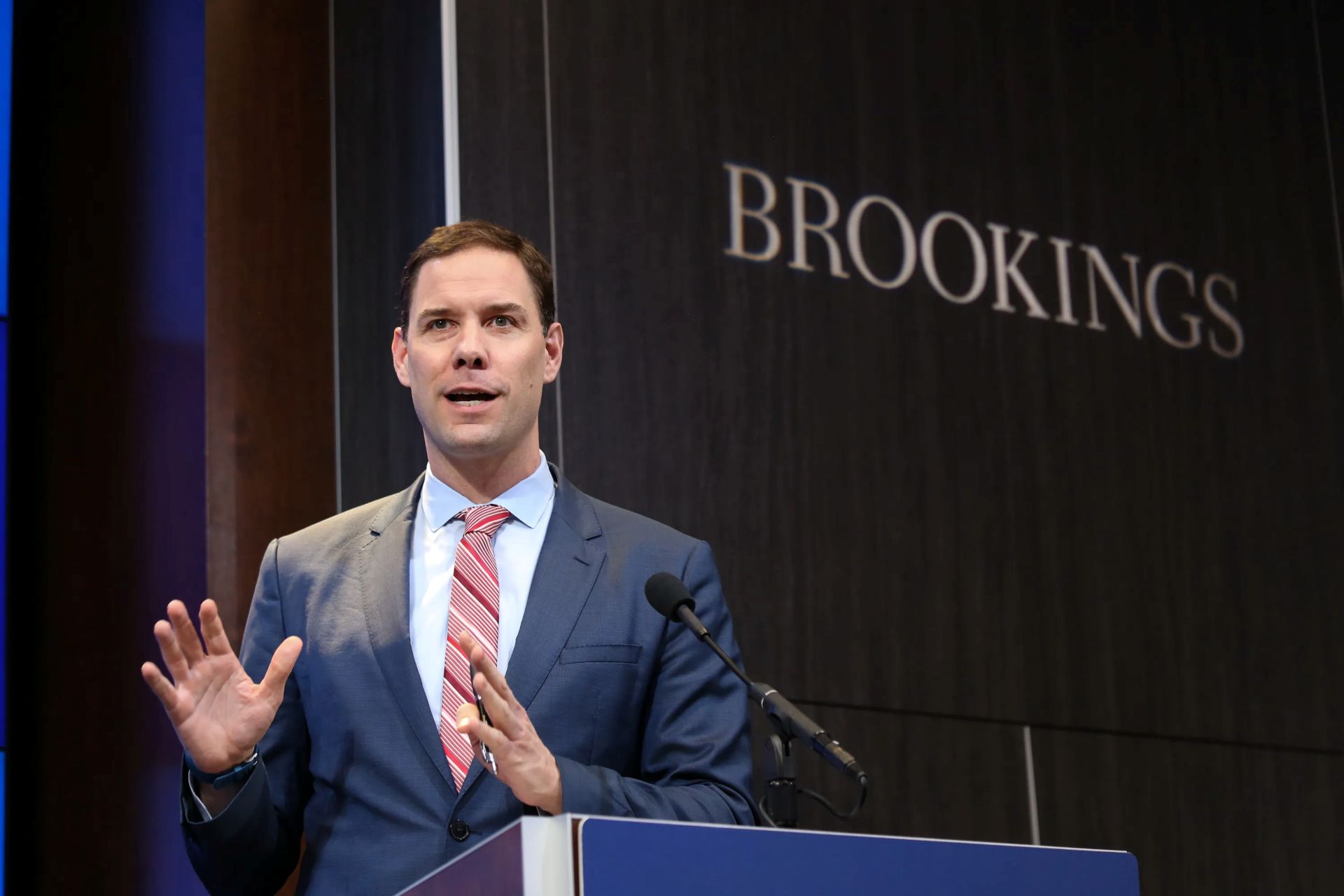 John McArthur speaking in front of a podium with Brookings in the background. 