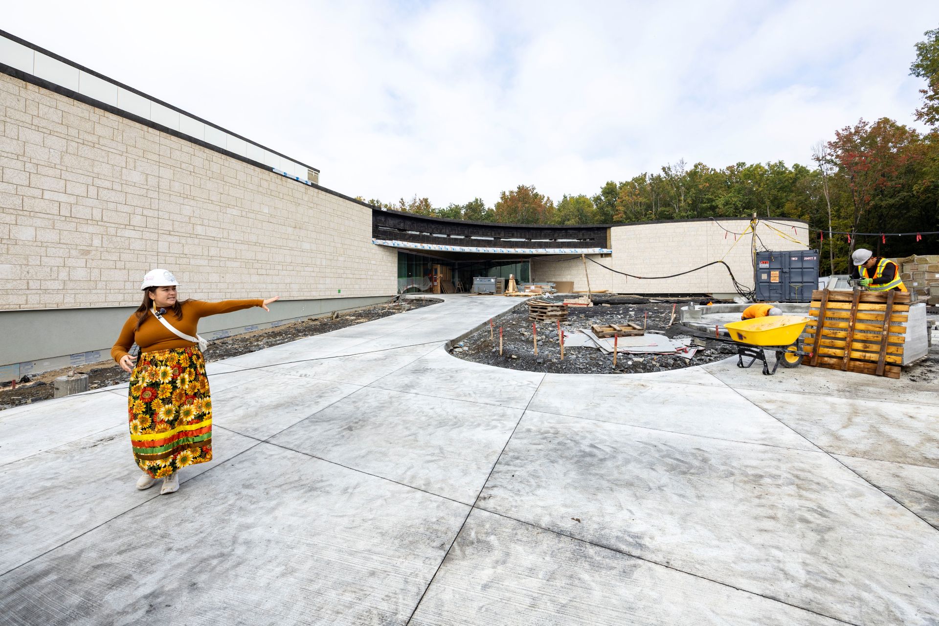 Cross stands outside a section of the building covered in off-white tiles. She is pointing to an area that has paving stones and will accommodate outside gatherings. 